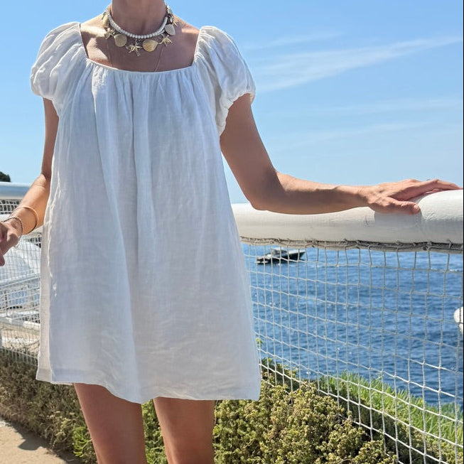 A woman wearing a white linen puff-sleeve mini dress with a square neckline and a chunky gold star necklace, standing outdoors on a sunny day next to the water.