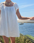 A woman wearing a white linen puff-sleeve mini dress with a square neckline and a chunky gold star necklace, standing outdoors on a sunny day next to the water.