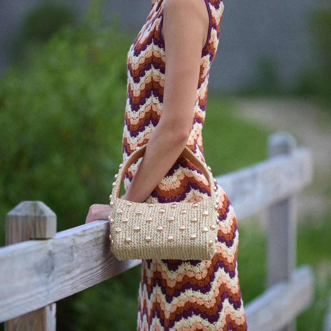 Person wearing a colorful dress holding The Lachlan raffia bag with gold stud accents